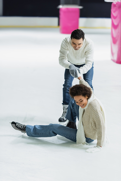 Smiling african american woman sitting on ice near boyfriend on rink