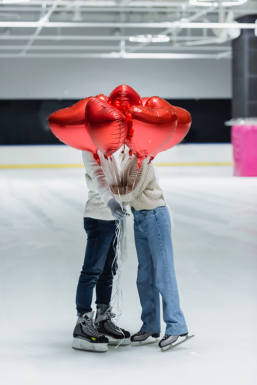 Young couple in warm clothes holding balloons in shape of heart on ice rink