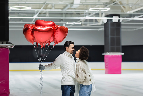 Side view of smiling african american woman hugging boyfriend with balloons in shape of heart on ice rink