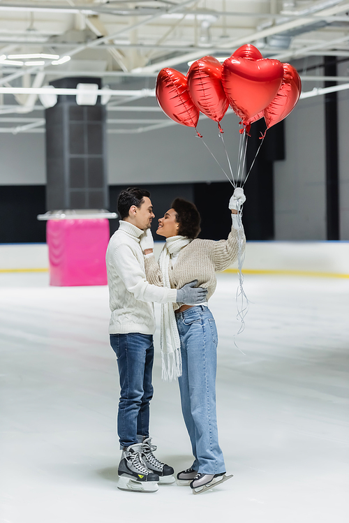 Side view of cheerful african american woman holding balloons in heart shape and touching boyfriend on ice rink