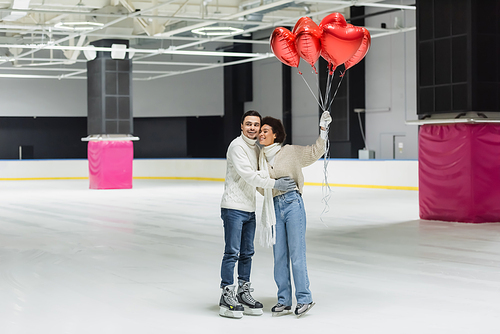 Positive interracial couple hugging and holding balloons in shape of heart on ice rink