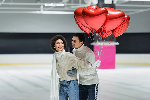 Smiling man in sweater hugging african american girlfriend with balloons in heart shape on ice rink