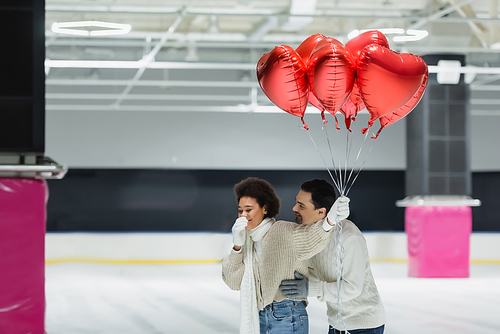 Young man hugging cheerful african american girlfriend with balloons in heart shape on ice rink