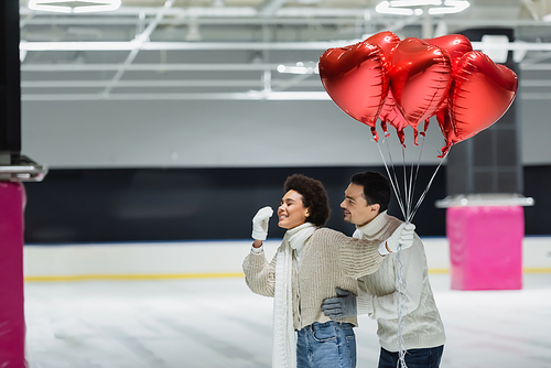 Young man hugging cheerful african american girlfriend in sweater holding red heart shaped balloons on ice rink