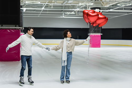 Side view of smiling interracial couple holding heart shaped balloons while ice skating on rink