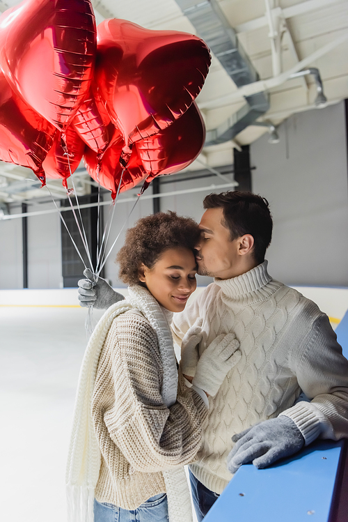 Young man in gloves and sweater kissing african american girlfriend and holding heart shaped balloons on ice rink