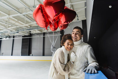 Cheerful interracial couple holding heart shaped balloons and looking at camera on ice rink