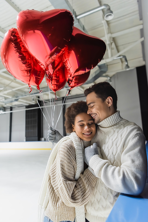 Young man in sweater hugging african american girlfriend and holding red heart shaped balloons on ice rink