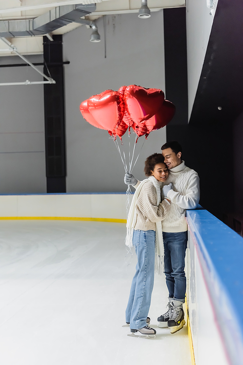 Cheerful interracial couple holding hands and balloons in shape of heart on ice rink