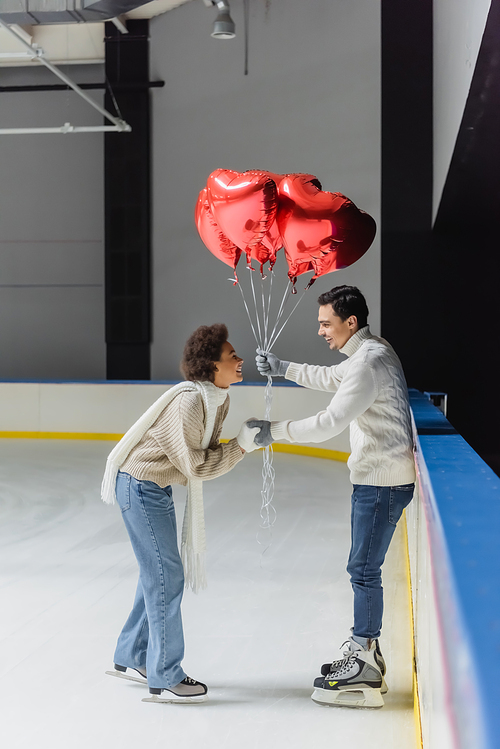 Side view of positive african american woman holding hand of boyfriend with heart shaped balloons on ice rink