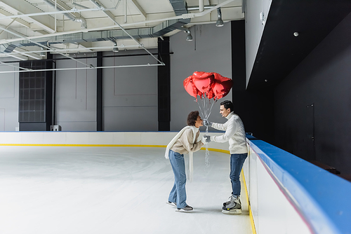 Side view of smiling man holding heart shaped balloons and hand of african american girlfriend on ice rink