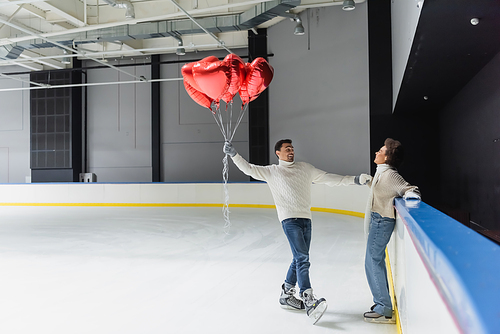 Cheerful african american woman holding hand of boyfriend with heart shaped balloons on ice rink