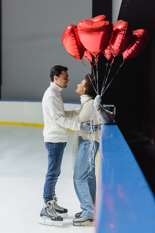 Side view of young man in ice skates hugging african american girlfriend holding heart shaped balloons on rink