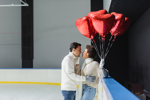 Side view of cheerful african american woman kissing boyfriend and holding heart shaped balloons on ice rink