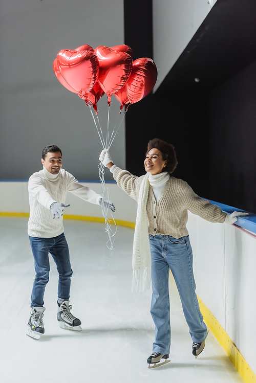 Smiling african american woman holding red heart shaped balloons while ice skating near boyfriend on rink