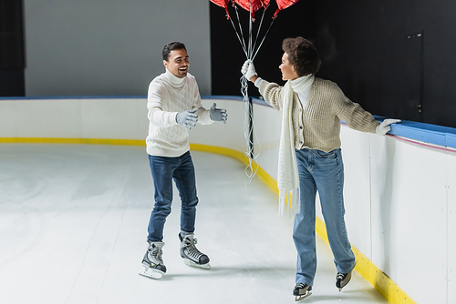 Smiling man ice skating near african american girlfriend with balloons on rink