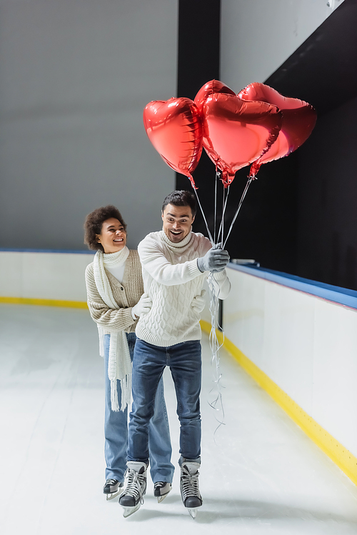 Positive multiethnic couple with heart shaped balloons ice skating on rink