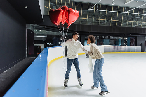 Positive interracial couple in warm sweaters holding heart shaped balloons while ice skating on rink