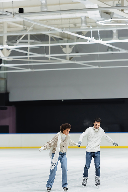 Smiling multiethnic couple in gloves and sweaters ice skating on rink