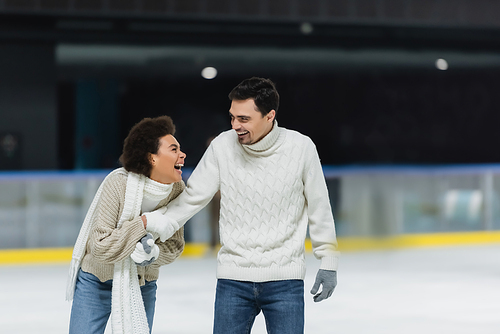 Cheerful african american woman holding hand of boyfriend in sweater and gloves on ice rink