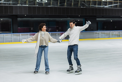 Positive multiethnic couple in gloves ice skating and having fun on rink