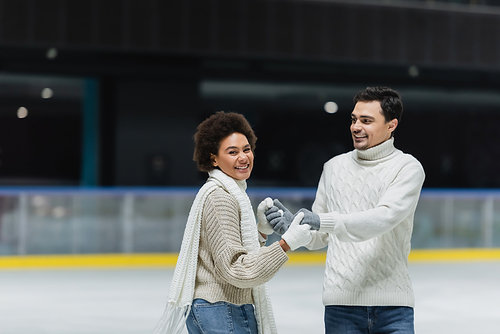 Cheerful african american woman looking at camera and holding hands of boyfriend on ice rink