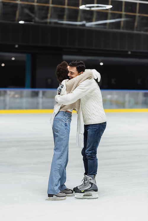 Smiling man in sweater embracing african american girlfriend on ice rink