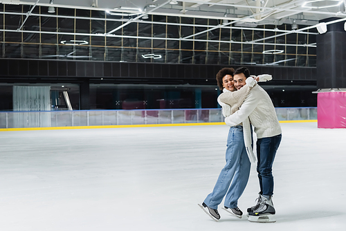 Multiethnic couple in warm clothes embracing and looking at camera on ice rink