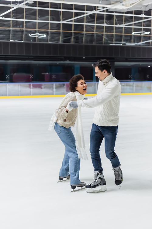 Excited african american woman holding hands of boyfriend while ice skating on rink