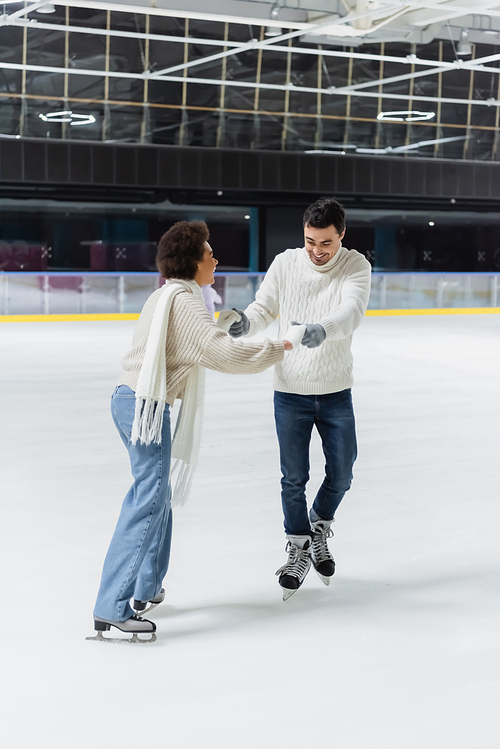 Cheerful interracial couple in jeans and sweaters holding hands while ice skating on rink