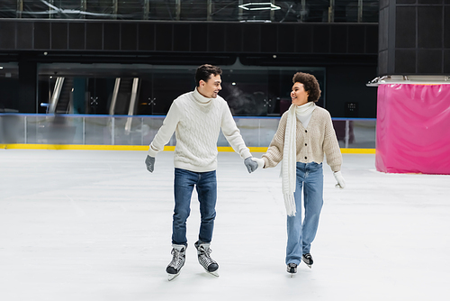 Cheerful interracial couple in gloves and sweaters skating on ice rink