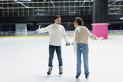 Smiling interracial couple in gloves and sweaters holding hands on ice rink