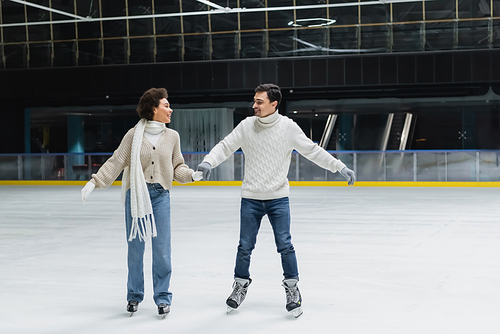 Smiling african american woman in jeans and sweater ice skating with boyfriend on rink