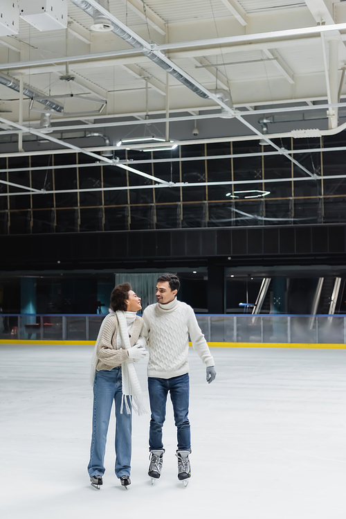 Young african american woman in sweater and jeans ice skating and looking at boyfriend on rink