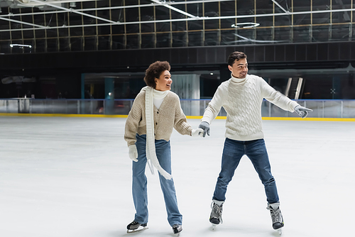 Young man pointing with finger and ice skating with african american girlfriend on rink