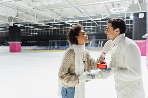 Side view of smiling man holding heart shaped gift box and hand of african american girlfriend on ice rink