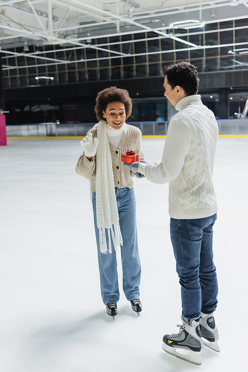 Smiling african american woman looking at heart shaped gift near boyfriend on ice rink