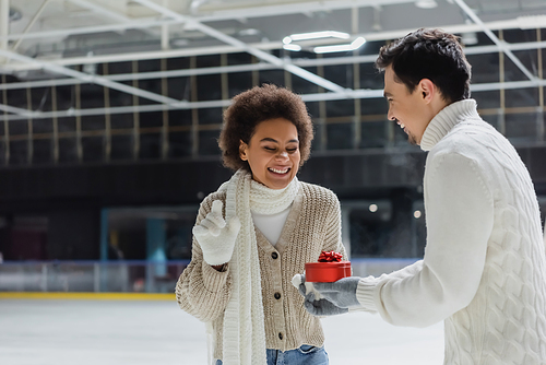 Cheerful man holding heart shaped gift near excited african american girlfriend on ice rink