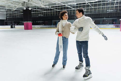 Smiling african american woman holding gift box and looking at boyfriend on ice rink