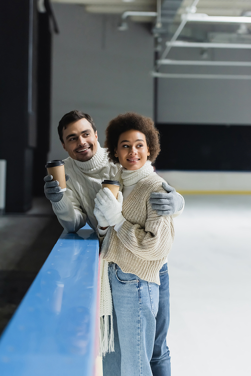 Smiling man hugging african american girlfriend with coffee to go on ice rink