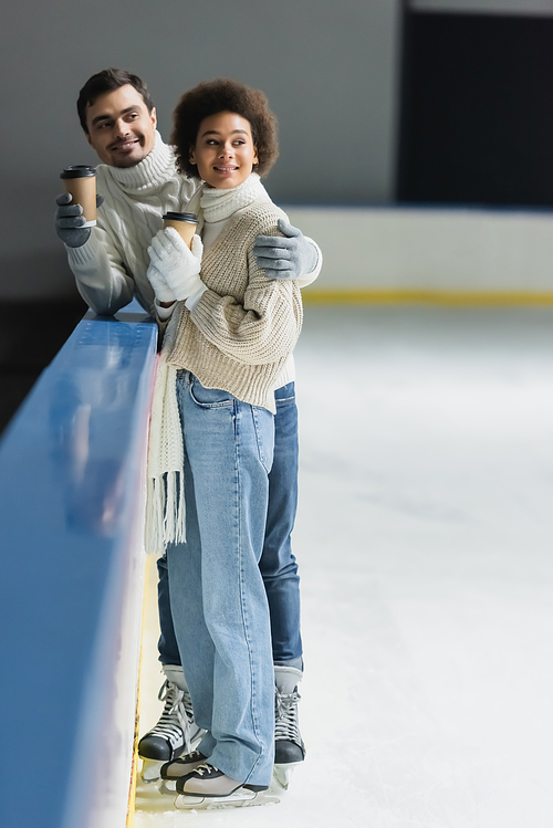 Smiling man hugging african american girlfriend with paper cup on ice rink