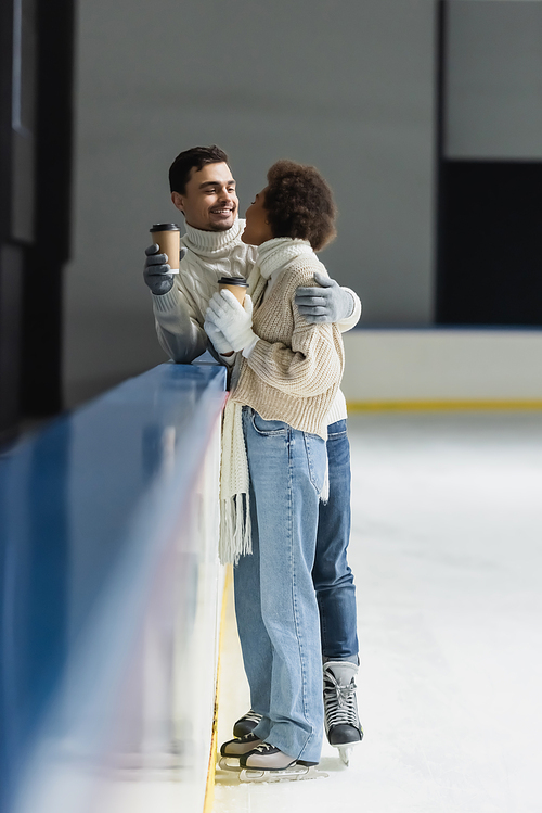 Young man in gloves holding coffee to go and hugging african american girlfriend on ice rink