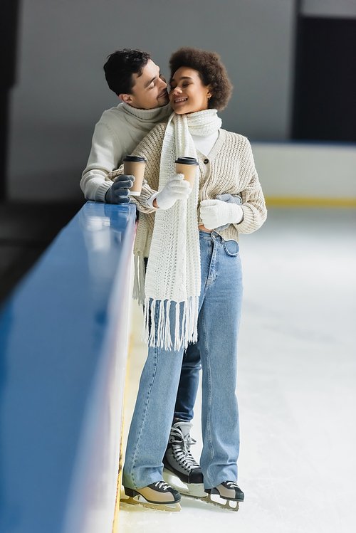 Smiling man kissing and hugging african american girlfriend with coffee to go on ice rink