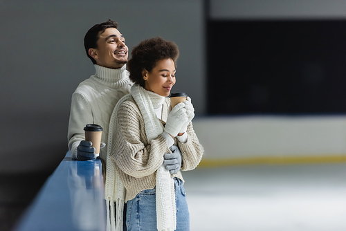 Positive interracial couple in gloves holding coffee to go on ice rink