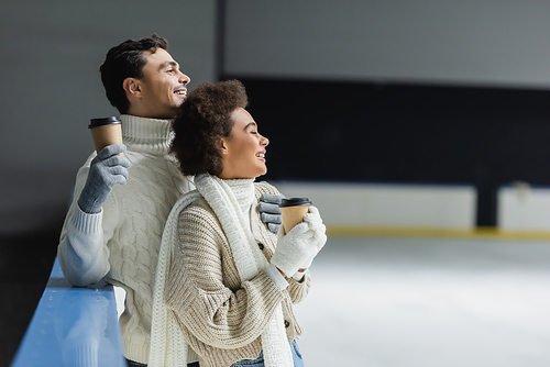 Side view of cheerful interracial couple holding coffee to go and looking away on ice rink