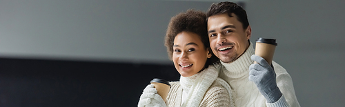 Positive multiethnic couple in warm clothes holding paper cups and looking at camera, banner