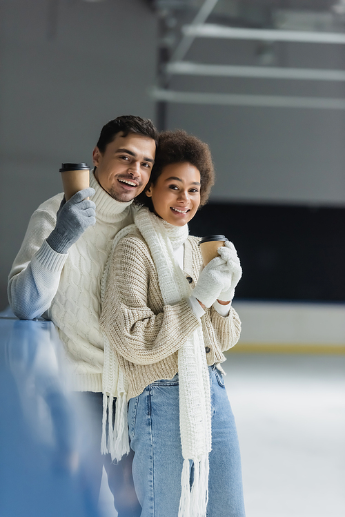 Cheerful interracial couple holding takeaway drink and looking at camera on ice rink