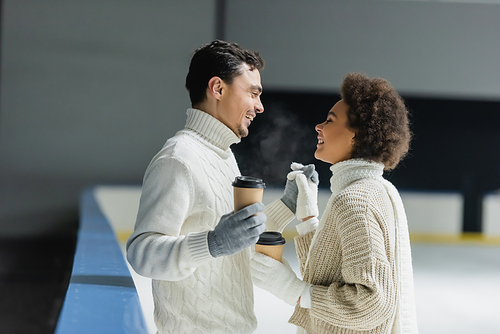 Side view of smiling interracial couple holding hands and coffee to go on ice rink