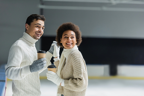 Positive multiethnic couple holding hands and coffee to go on ice rink