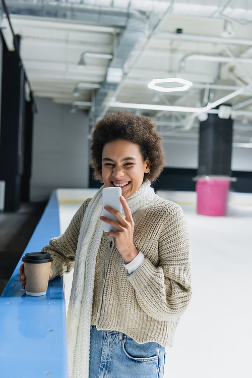 Smiling african american woman holding smartphone and paper cup on ice rink